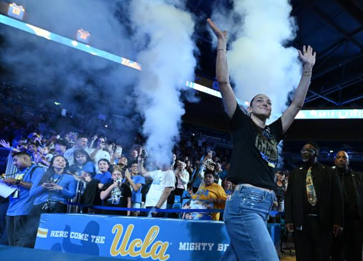 A UCLA fan raises both arms in celebration as blue and white smoke fills the arena. Behind them, a cheering crowd gathers near a barrier displaying UCLA branding.