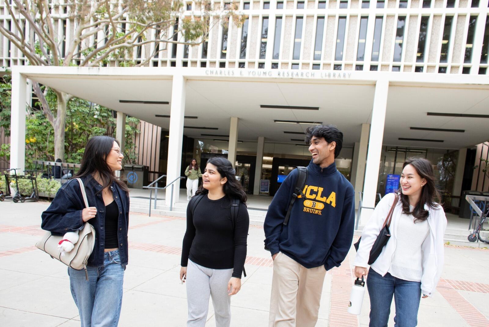 Four UCLA students walk together outside Charles E. Young Research Library, carrying backpacks and chatting on campus.